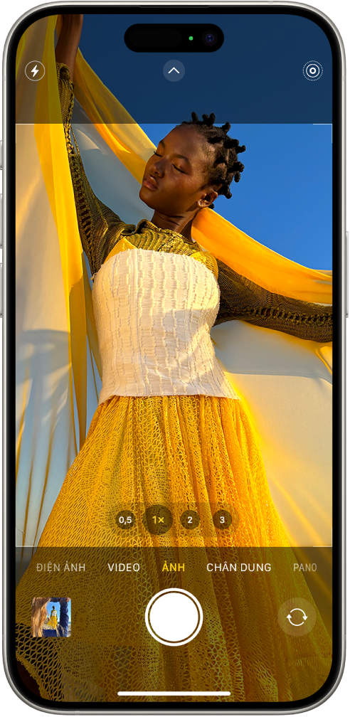 Camera ở chế độ Ảnh, với các chế độ khác ở bên trái và phải bên dưới kính ngắm. Các nút cho Flash, Điều khiển camera và Live Photos xuất hiện ở đầu màn hình. Nút Trình xem ảnh và video ở góc dưới cùng bên trái. Nút Chụp ảnh ở dưới cùng ở giữa và nút Chọn camera mặt sau ở góc dưới cùng bên phải.